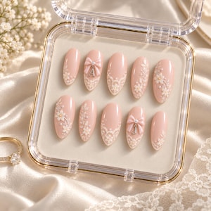 May include: A set of ten almond-shaped press-on nails in a soft blush pink hue, featuring intricate white lace patterns, raised floral embellishments, small pearl accents, and two nails adorned with 3D bows and silver letter charms A and J.