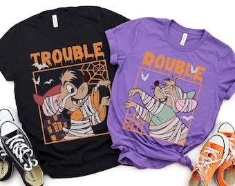 May include: Two black and purple t-shirts with a cartoon image of a mummified mouse. The black shirt says "Trouble" and the purple shirt says "Double".