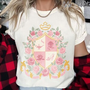 Puede incluir: Camiseta blanca con un dise&ntilde;o de escudo rosa y dorado con una corona, rosas e iconos de cuentos de hadas. El escudo est&aacute; rodeado de rosas y cintas rosas. El texto "Color: White" es visible en la esquina superior derecha.