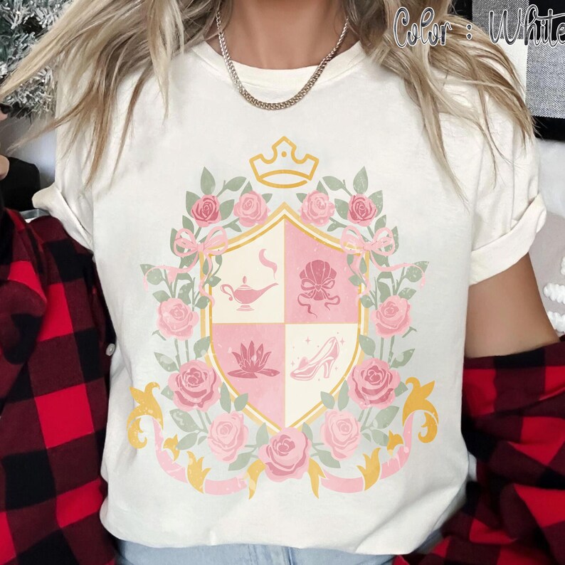 Puede incluir: Camiseta blanca con un dise&ntilde;o de escudo rosa y dorado con una corona, rosas e iconos de cuentos de hadas. El escudo est&aacute; rodeado de rosas y cintas rosas. El texto "Color: White" es visible en la esquina superior derecha.