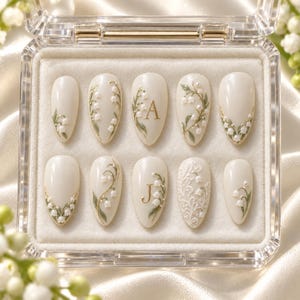 May include: A set of ten almond-shaped press-on nails in a clear display case. The nails are cream-colored and feature delicate hand-painted lily of the valley floral designs in green and white, with gold letter accents A and J on two nails.