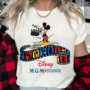 Pu&ograve; includere: T-shirt bianca con Topolino che tiene una clapperboard con la scritta "ACTION". Il design include una bobina di film e una striscia di film colorata con personaggi Disney. I loghi Disney e MGM Studios sono stampati sotto.