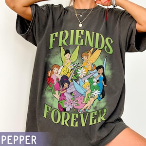 May include: Dark gray t-shirt featuring a graphic of several fairies with colorful wings and floral accents. The words "FRIENDS FOREVER" are printed in green. The shirt has a relaxed fit.