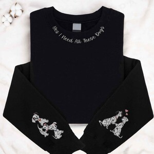 Op de afbeelding: Zwart sweatshirt met de tekst "Ja, ik heb al deze honden nodig" geborduurd op de halslijn. De mouwen hebben geborduurde afbeeldingen van Dalmati&euml;rs.