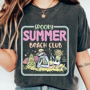 May include: Dark gray t-shirt featuring a graphic with the text "Spooky Summer Beach Club." The design includes illustrations of characters from "The Nightmare Before Christmas" in a beach setting, with a pumpkin and Jack Skellington.
