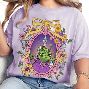 May include: Purple t-shirt with a cartoon image of a green chameleon wearing a purple dress and a crown. The chameleon is surrounded by flowers and lanterns. The background is a purple and blue gradient.