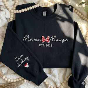 May include: A black crewneck sweatshirt featuring white embroidered text that reads Mama Mouse with a red and white polka dot bow icon in the center and EST. 2018 below it. The left sleeve features the embroidered names Zane and Zachary.