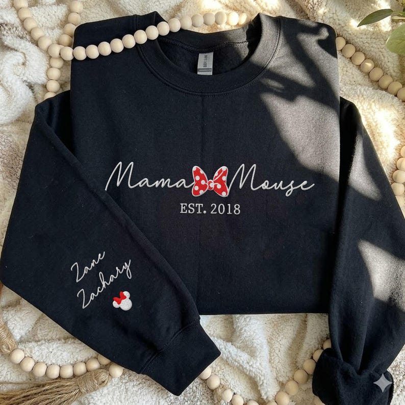 May include: A black crewneck sweatshirt featuring white embroidered text that reads Mama Mouse with a red and white polka dot bow icon in the center and EST. 2018 below it. The left sleeve features the embroidered names Zane and Zachary.
