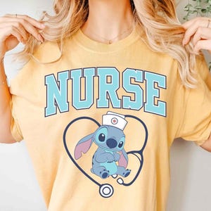 May include: Yellow t-shirt with the word "NURSE" in light blue block letters. Below the word is a blue cartoon character wearing a nurse's hat, inside a blue heart-shaped stethoscope.