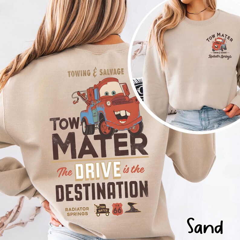 May include: Sand-colored sweatshirt featuring a cartoon image of Tow Mater, a red and blue tow truck, with the text "Towing & Salvage" and "Tow Mater". The phrase "The Drive is the Destination" is below the graphic. The sweatshirt also has the text "Radiator Springs".