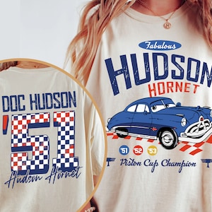 Könnte beinhalten: Zwei beige T-Shirts mit einer blauen Autografik und Text. Das vordere Shirt hat den Text "Fabulous Hudson Hornet" und das hintere Shirt hat den Text "Doc Hudson '51 Hudson Hornet".