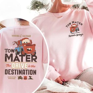 May include: A light pink sweatshirt featuring a cartoon image of Tow Mater from the movie Cars. The front of the sweatshirt has the text "TOW MATER" and "The DRIVE is the DESTINATION". The back of the sweatshirt has the text "TOWING & SALVAGE".