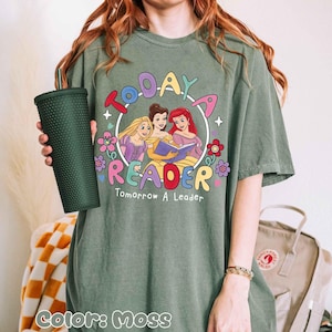 May include: Moss green t-shirt featuring a colorful graphic of three cartoon princesses reading a book, surrounded by flowers and the words "Today a Reader, Tomorrow a Leader." A dark green tumbler is held in the left hand.