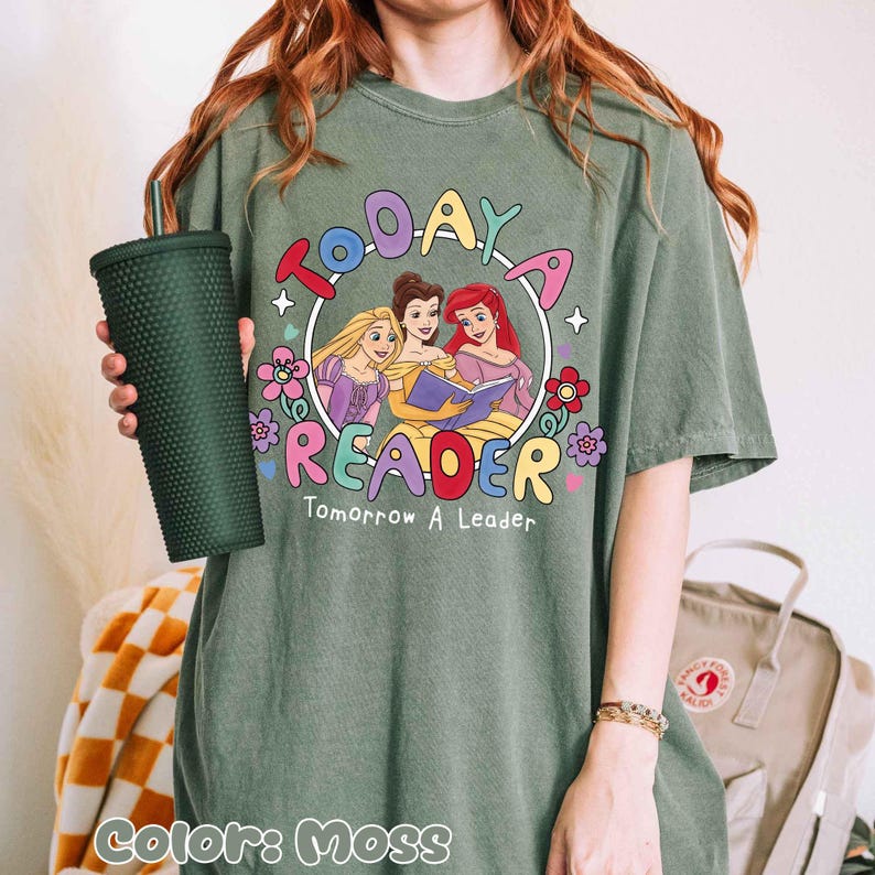 May include: Moss green t-shirt featuring a colorful graphic of three cartoon princesses reading a book, surrounded by flowers and the words "Today a Reader, Tomorrow a Leader." A dark green tumbler is held in the left hand.