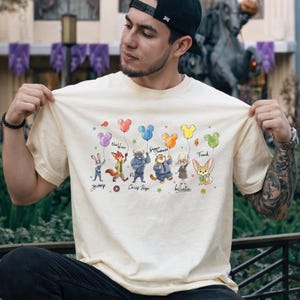 Op de afbeelding: Een cr&egrave;mekleurig t-shirt met stripfiguren die Mickey Mouse-vormige ballonnen vasthouden. De personages zijn onder meer een konijn, een vos, een stier, een tijger en een konijn. Het shirt heeft de namen van de personages eronder geschreven.