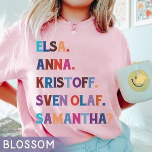 以下が含まれることがあります： エルサ、アナ、クリストフ、スヴェン・オラフ、サマンサ？という名前が様々な色でプリントされた、薄いピンク色のスウェットシャツ。「BLOSSOM」の文字が下部にプリントされています。黄色いスマイリーフェイスのマグカップを持っています。