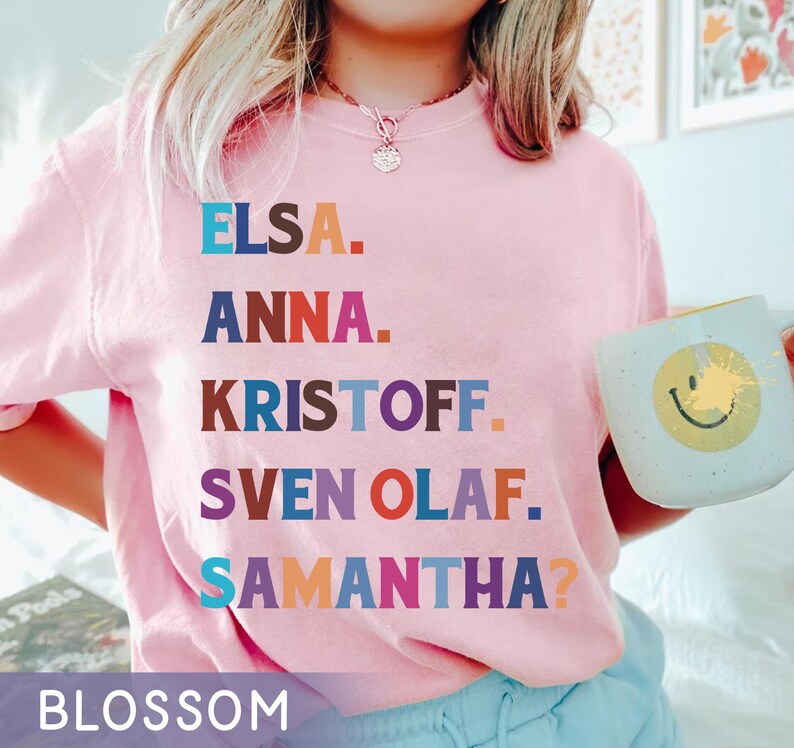 以下が含まれることがあります： エルサ、アナ、クリストフ、スヴェン・オラフ、サマンサ？という名前が様々な色でプリントされた、薄いピンク色のスウェットシャツ。「BLOSSOM」の文字が下部にプリントされています。黄色いスマイリーフェイスのマグカップを持っています。
