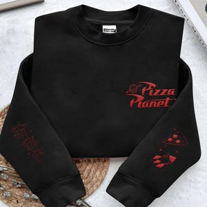 May include: A black long-sleeve sweatshirt featuring red embroidered designs. The chest displays the text Pizza Planet with a planet icon. The left sleeve shows three small alien figures, while the right sleeve features a pizza slice and a rocket.