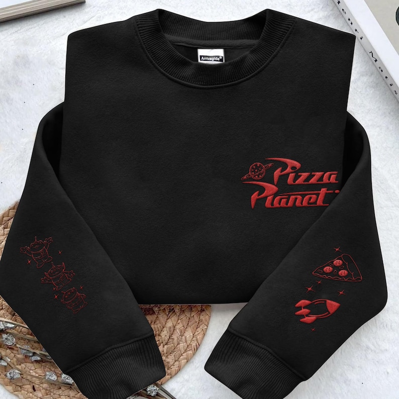 May include: A black long-sleeve sweatshirt featuring red embroidered designs. The chest displays the text Pizza Planet with a planet icon. The left sleeve shows three small alien figures, while the right sleeve features a pizza slice and a rocket.