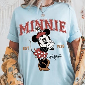 May include: Light blue t-shirt featuring a graphic of Minnie Mouse. The design includes the word "MINNIE" in red block letters, the year "1928", and the word "Minnie" in script. Minnie Mouse is wearing a red polka dot dress and hat.