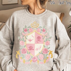 Puede incluir: Sudadera gris deportiva con un dise&ntilde;o de escudo rosa y blanco con una corona, rosas y lazos. El escudo incluye una l&aacute;mpara, una concha, una flor de loto y un tac&oacute;n alto. El texto "Color: Sport Grey" est&aacute; impreso en la parte superior.