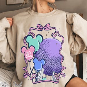 May include: Beige sweatshirt featuring a colorful graphic of a purple sphere, three Mickey Mouse-shaped balloons in blue, pink, and teal, and a pink ribbon. The design includes a bow and small stars.