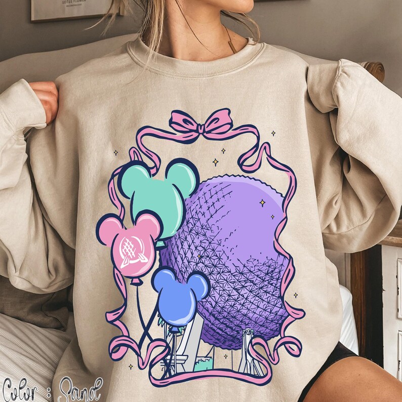 May include: Beige sweatshirt featuring a colorful graphic of a purple sphere, three Mickey Mouse-shaped balloons in blue, pink, and teal, and a pink ribbon. The design includes a bow and small stars.