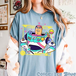 May include: Ice blue t-shirt featuring a cartoon illustration of Buzz Lightyear in his spacesuit. The design includes a yellow frame, pink stars, and a smiling emoji. The text "Color: Ice Blue" is at the bottom.