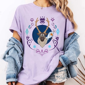 Puede incluir: Camiseta lila claro con un dise&ntilde;o de dibujos animados de Sven el reno y Olaf el mu&ntilde;eco de nieve de la pel&iacute;cula Frozen. El dise&ntilde;o est&aacute; enmarcado por un borde decorativo morado y azul. Las mangas est&aacute;n dobladas.