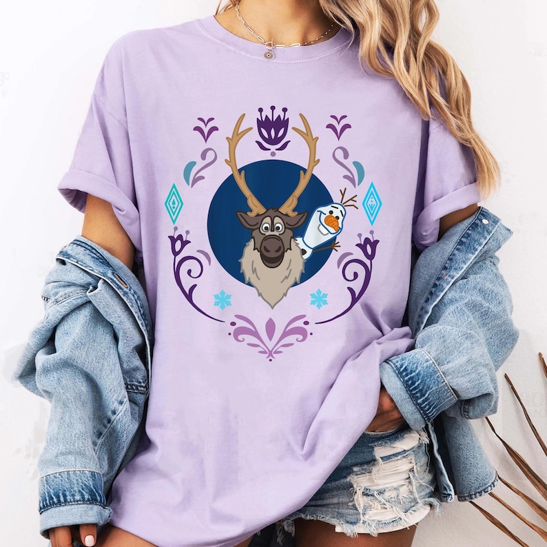 Puede incluir: Camiseta lila claro con un dise&ntilde;o de dibujos animados de Sven el reno y Olaf el mu&ntilde;eco de nieve de la pel&iacute;cula Frozen. El dise&ntilde;o est&aacute; enmarcado por un borde decorativo morado y azul. Las mangas est&aacute;n dobladas.