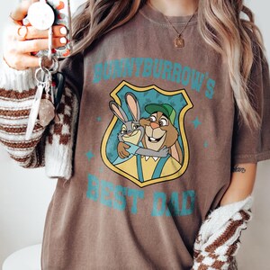 Puede incluir: Camiseta marr&oacute;n con un dibujo animado de dos conejos abraz&aacute;ndose y el texto turquesa "BUNNYBURROW'S BEST DAD". El dibujo est&aacute; dentro de un escudo. La camiseta tiene un aspecto vintage y desgastado.