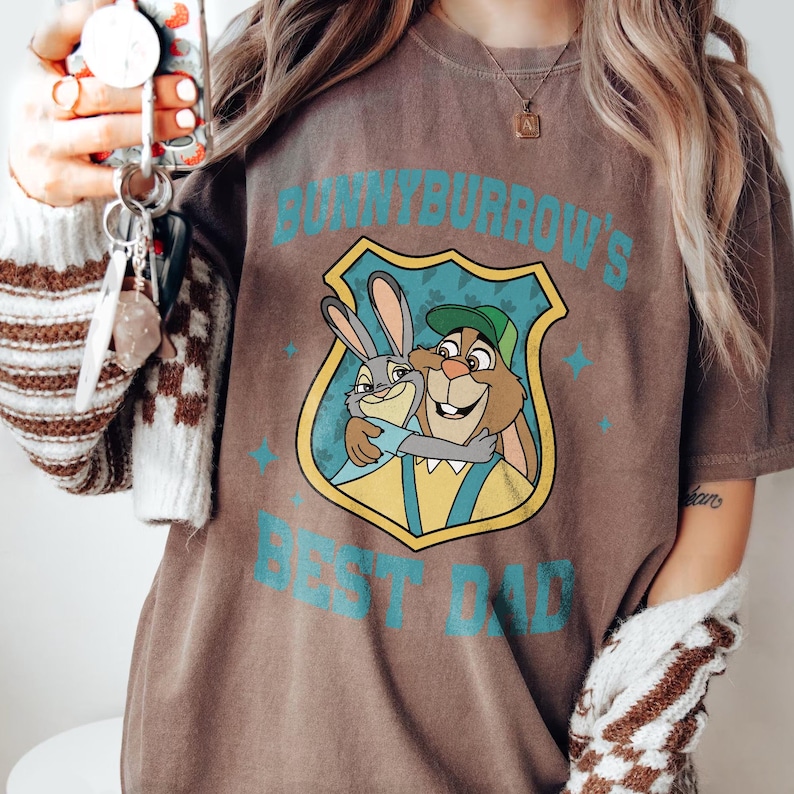 Puede incluir: Camiseta marr&oacute;n con un dibujo animado de dos conejos abraz&aacute;ndose y el texto turquesa "BUNNYBURROW'S BEST DAD". El dibujo est&aacute; dentro de un escudo. La camiseta tiene un aspecto vintage y desgastado.