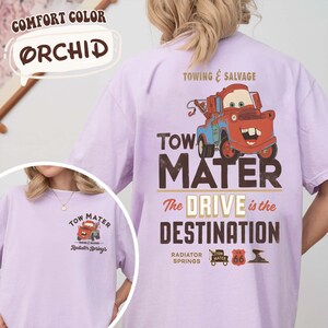 May include: Orchid-colored t-shirt featuring a graphic of Tow Mater from the movie Cars. The back of the shirt displays "Towing & Salvage" and "The Drive is the Destination." The front has a smaller graphic. Made of Comfort Color material.