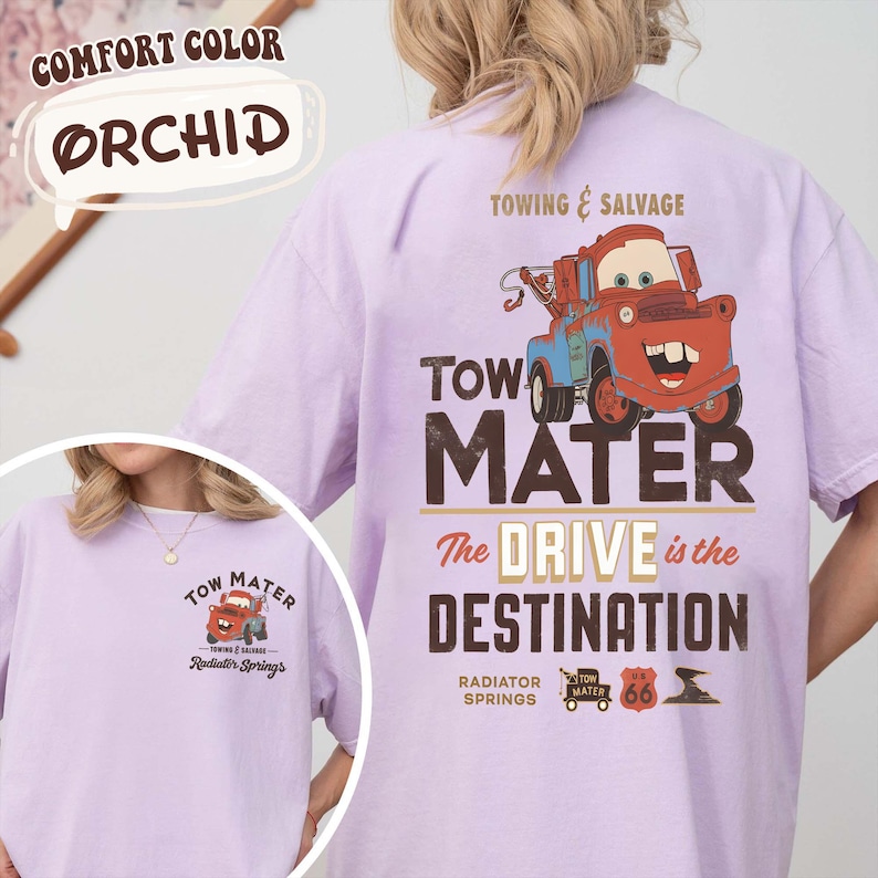 May include: Orchid-colored t-shirt featuring a graphic of Tow Mater from the movie Cars. The back of the shirt displays "Towing & Salvage" and "The Drive is the Destination." The front has a smaller graphic. Made of Comfort Color material.