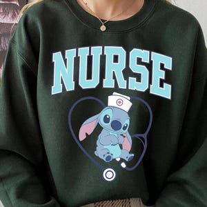 May include: Dark green sweatshirt with the word "NURSE" in light blue block letters. Below the word is a cartoon character wearing a nurse's hat inside a blue heart-shaped stethoscope.