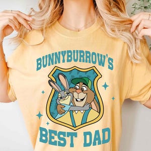 Puede incluir: Camiseta amarilla clara con un gr&aacute;fico de dos conejos de dibujos animados abraz&aacute;ndose dentro de un escudo. El escudo tiene el texto "BUNNYBURROW'S BEST DAD" en azul.