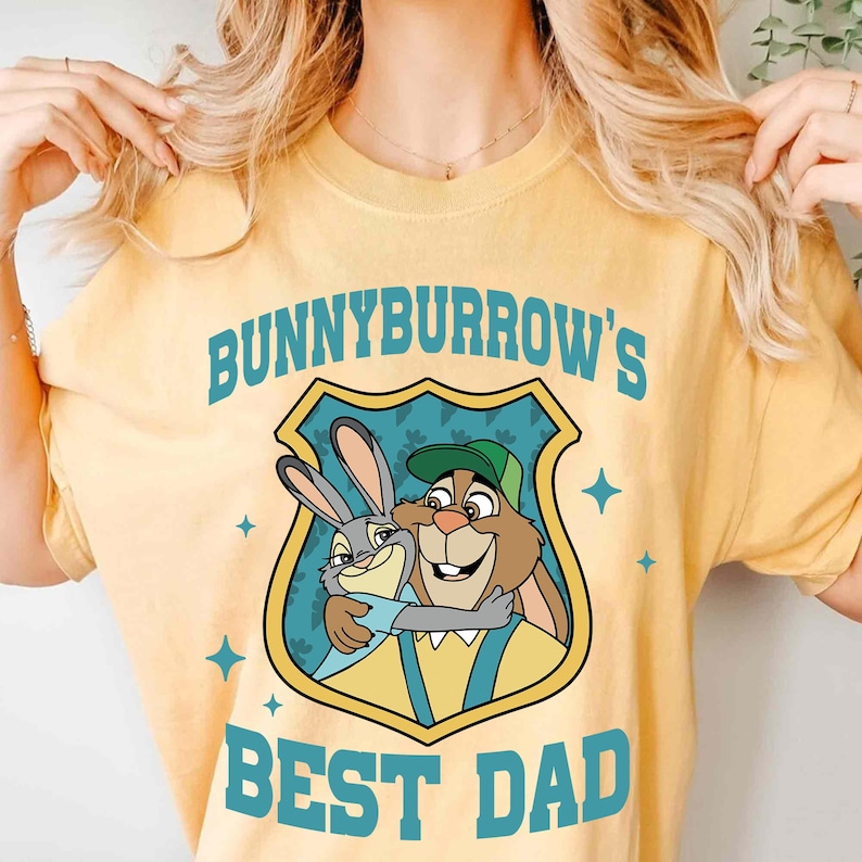 Puede incluir: Camiseta amarilla clara con un gr&aacute;fico de dos conejos de dibujos animados abraz&aacute;ndose dentro de un escudo. El escudo tiene el texto "BUNNYBURROW'S BEST DAD" en azul.