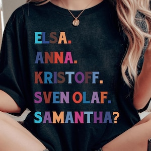 以下が含まれることがあります： エルサ、アナ、クリストフ、スヴェン、オラフ、サマンサ？という名前がカラフルなブロックレターでプリントされた黒いTシャツ。Tシャツはクルーネックと半袖です。テキストは遊び心のある漫画のようなフォントです。