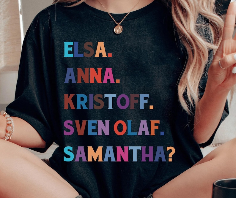 以下が含まれることがあります： エルサ、アナ、クリストフ、スヴェン、オラフ、サマンサ？という名前がカラフルなブロックレターでプリントされた黒いTシャツ。Tシャツはクルーネックと半袖です。テキストは遊び心のある漫画のようなフォントです。