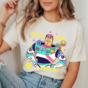 May include: Cream-colored t-shirt featuring a colorful cartoon illustration of Buzz Lightyear in a yellow outlined box with smiley faces and stars. The character is wearing a space suit with blue, teal, pink, and purple accents.