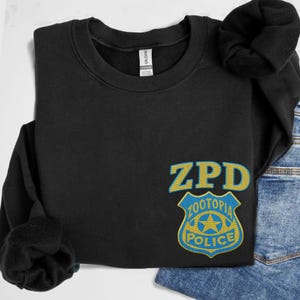 May include: A black crewneck sweatshirt featuring an embroidered ZPD logo and a Zootopia Police badge in yellow and blue thread on the lower right side. The garment has a ribbed collar and long sleeves with a soft fleece interior lining.