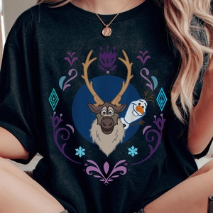 Puede incluir: Camiseta gris oscuro con una ilustraci&oacute;n de dibujos animados de Sven el reno y Olaf el mu&ntilde;eco de nieve de la pel&iacute;cula Frozen. El dise&ntilde;o est&aacute; enmarcado en un marco decorativo p&uacute;rpura y turquesa con copos de nieve y diamantes.