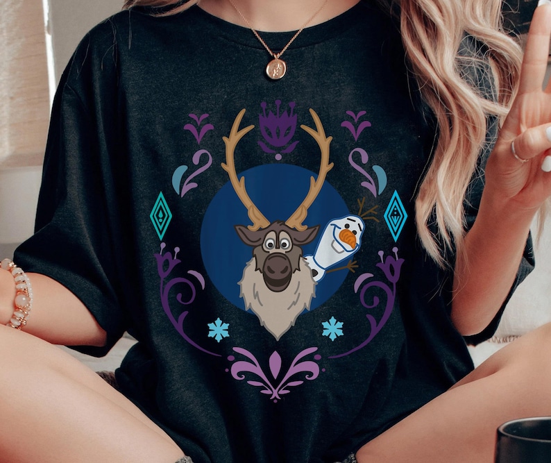 Puede incluir: Camiseta gris oscuro con una ilustraci&oacute;n de dibujos animados de Sven el reno y Olaf el mu&ntilde;eco de nieve de la pel&iacute;cula Frozen. El dise&ntilde;o est&aacute; enmarcado en un marco decorativo p&uacute;rpura y turquesa con copos de nieve y diamantes.