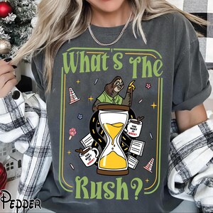 Puede incluir: Camiseta gris oscuro con un dise&ntilde;o gr&aacute;fico humor&iacute;stico. Presenta un perezoso, un reloj de arena y la frase "What's The Rush?". El dise&ntilde;o incluye tazas de caf&eacute; y elementos de carretera, con el texto "You want it when?" en las tazas.