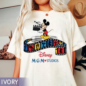 Pu&ograve; includere: T-shirt color avorio con un design Disney MGM Studios. La grafica presenta Mickey Mouse con una ciak, in piedi su una bobina di film, con una striscia di pellicola colorata sottostante. Il testo "Disney MGM Studios" &egrave; stampato in rosso e blu.