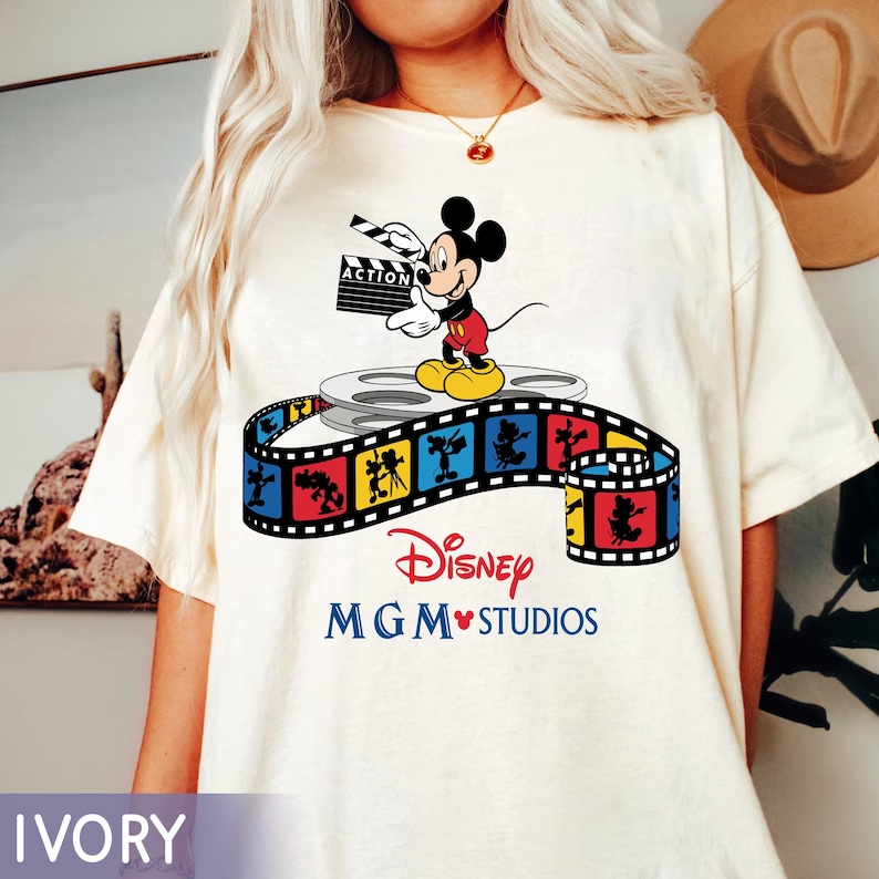 Pu&ograve; includere: T-shirt color avorio con un design Disney MGM Studios. La grafica presenta Mickey Mouse con una ciak, in piedi su una bobina di film, con una striscia di pellicola colorata sottostante. Il testo "Disney MGM Studios" &egrave; stampato in rosso e blu.