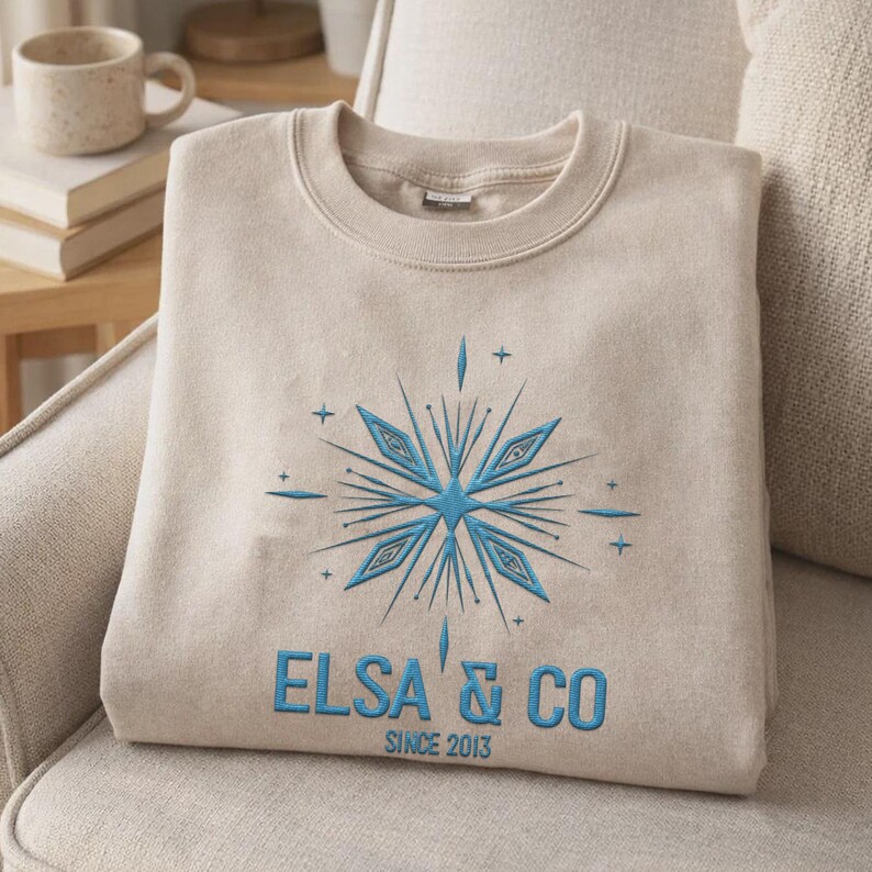 May include: A beige crewneck sweatshirt featuring an embroidered blue snowflake design centered on the chest. Below the snowflake, the text reads ELSA & CO in blue capital letters, with the smaller text SINCE 2013 printed underneath.