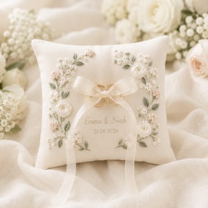 May include: A square off-white ring pillow featuring embroidered floral wreaths in shades of white, pink, and sage green. Two gold rings are tied to the center with a sheer ribbon. The text "Emma & Noah" and "21.09.2026" is embroidered below.