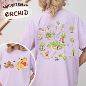 May include: Orchid-colored t-shirt featuring a map design on the back and a cartoon character on the front. The map design includes trees, paths, and text. The front of the shirt shows a cartoon character with honey pots and bees. The shirt is a Comfort Colors brand.