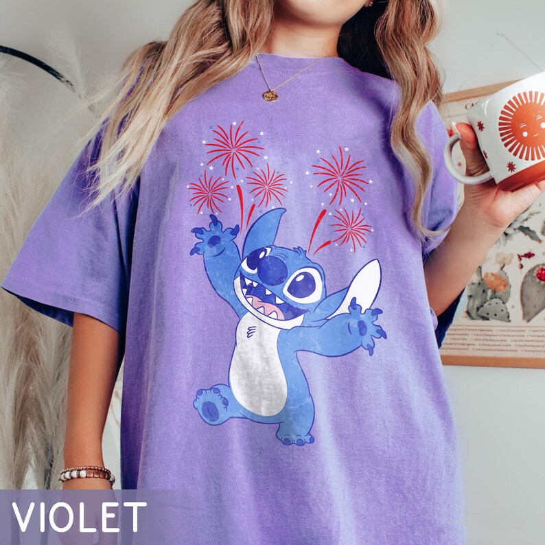 May include: Violet-colored t-shirt featuring a cartoon character with a blue body, white belly, and large eyes, with red fireworks. The character is smiling and has its arms outstretched. The word "VIOLET" is at the bottom of the shirt.